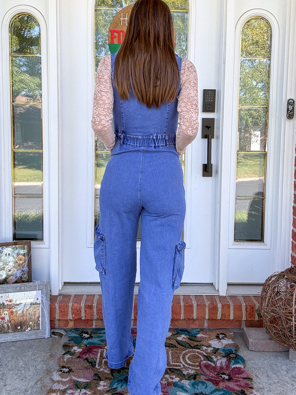 A woman stands with her back to the camera on a front porch, wearing a blue denim overall jumpsuit over a light pink lace long-sleeve top. The setting features a white double door with brick columns and a floral rug.