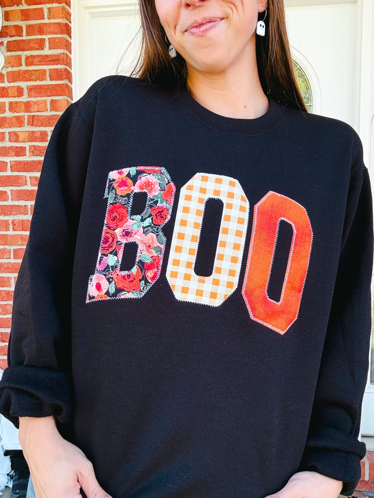 A woman wearing a black crewneck sweatshirt with large 'BOO' letter graphics. The letter 'B' has a red and pink floral pattern, 'O' has a white and orange gingham check pattern, and 'O' has a textured orange gradient.