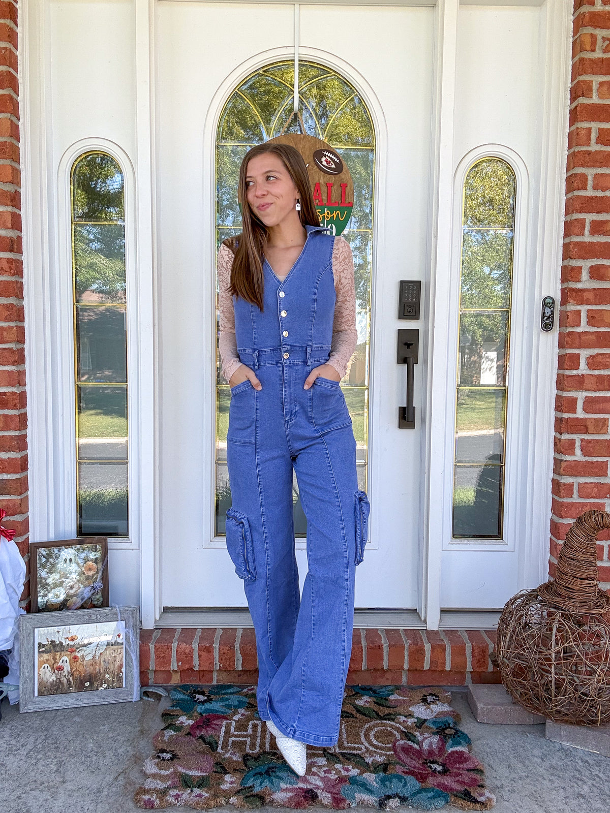 A woman stands on a front porch wearing a blue denim overall jumpsuit over a sheer beige lace long-sleeve top. The background shows a white front door with brick columns and a decorative doormat.