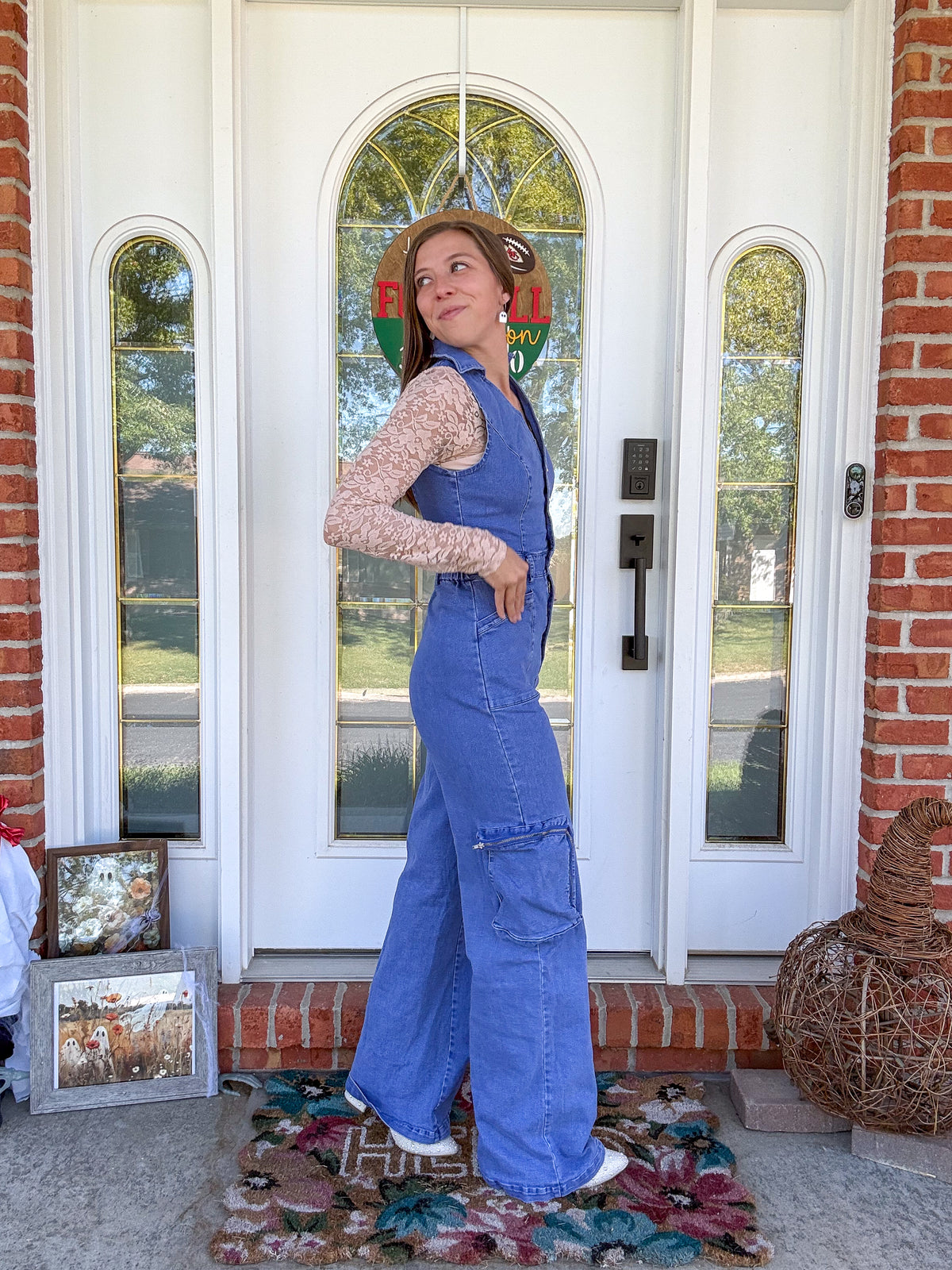 A woman wearing a blue denim overall jumpsuit over a beige lace long-sleeve top stands on a front porch in front of a white double door with brick columns.