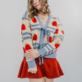 Model wearing "The Perfect Pick Cardigan" with a red skirt and holding her left arm over her right arm in a pose. The sweater is blue striped with strawberries covering it and two bows to tie it on.
