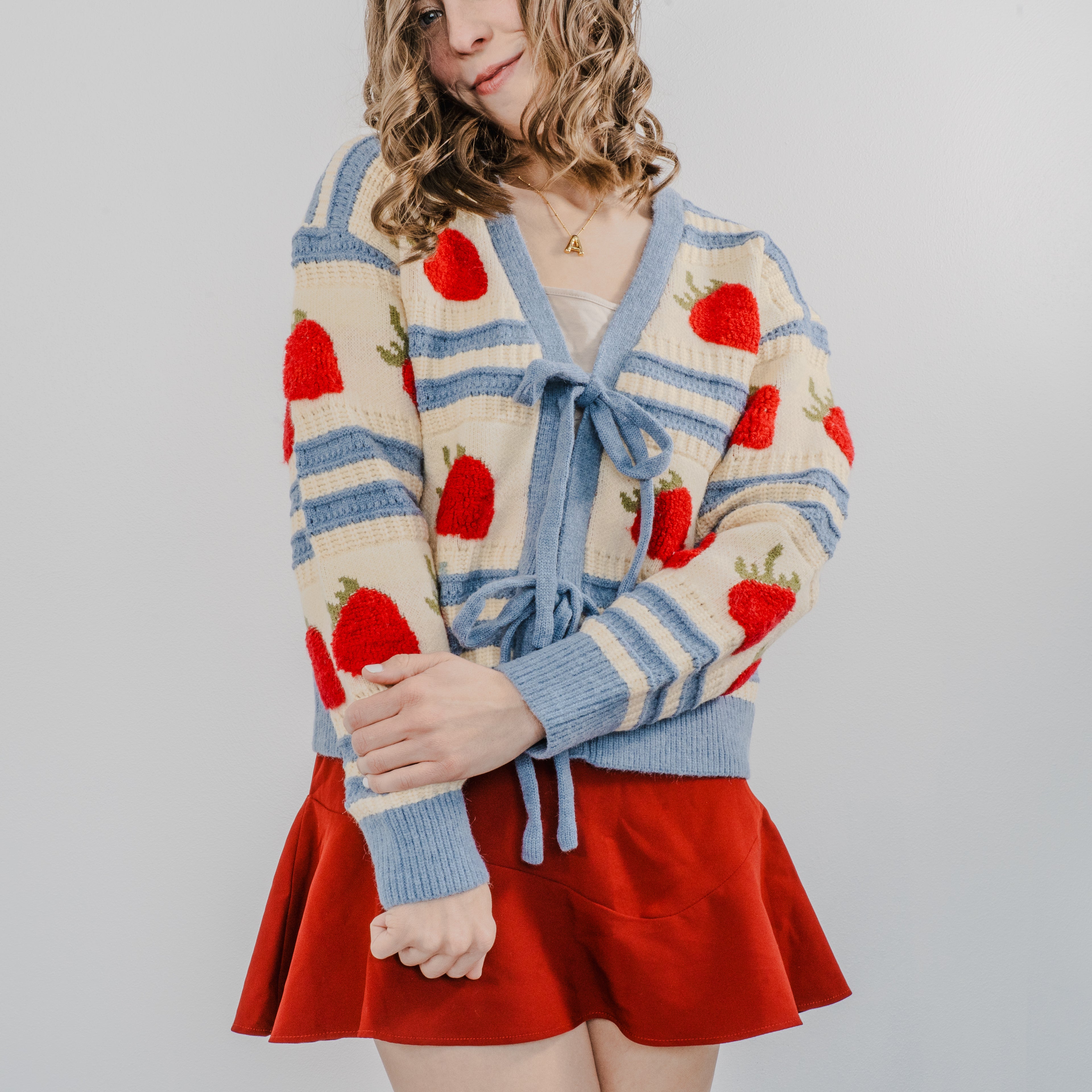 Model wearing "The Perfect Pick Cardigan" with a red skirt and holding her left arm over her right arm in a pose. The sweater is blue striped with strawberries covering it and two bows to tie it on.