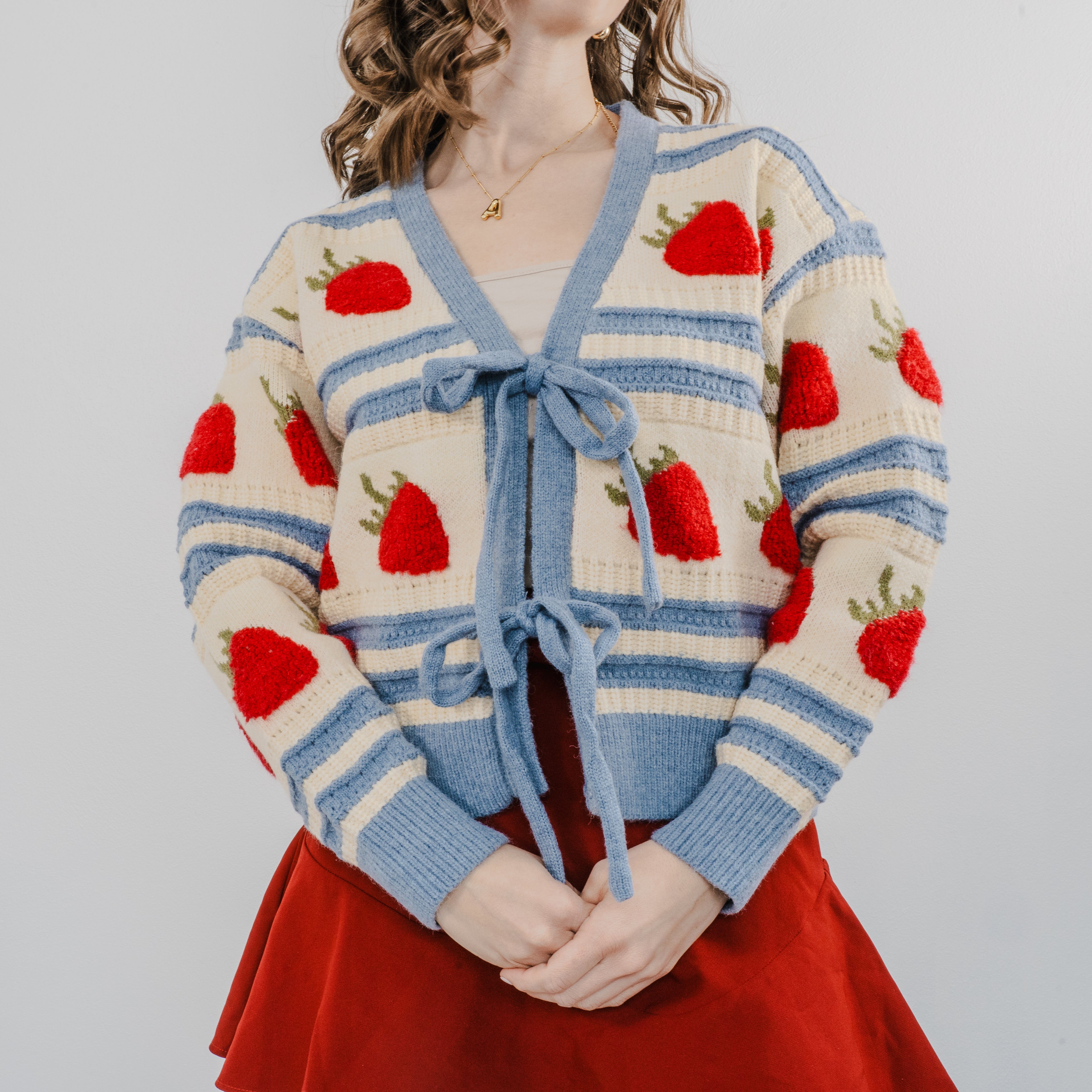 Up close shot of model wearing "The Perfect Pick Cardigan" with a red skirt. The sweater is blue striped with strawberries covering it and two bows to tie it on.