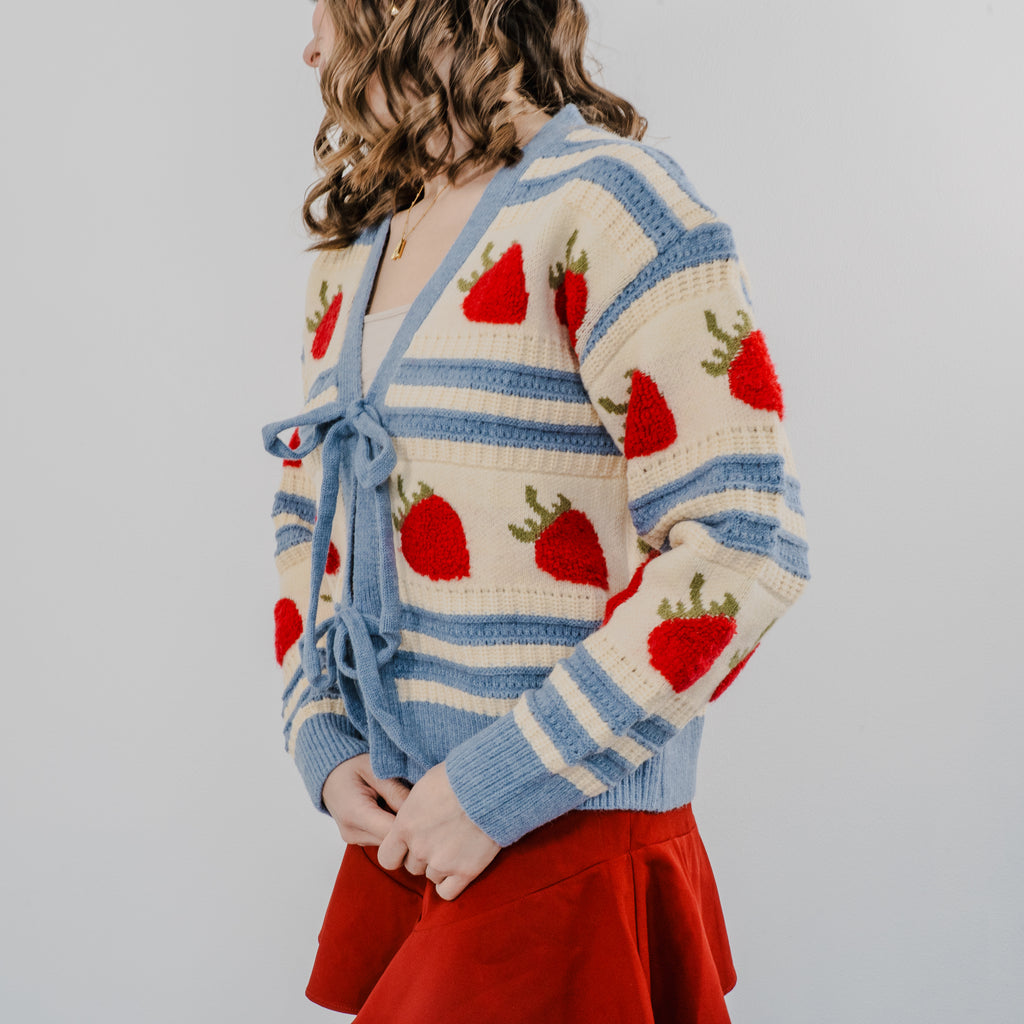 Side view of model wearing "The Perfect Pick Cardigan" with a red skirt. The sweater is blue striped with strawberries covering it and two bows to tie it on.