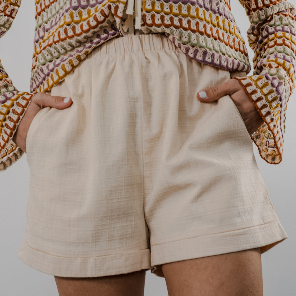 High-waisted cream crochet shorts with a functional drawstring on either side of hip to adjust waist. Model's hands are partially in pockets to show item has pockets.