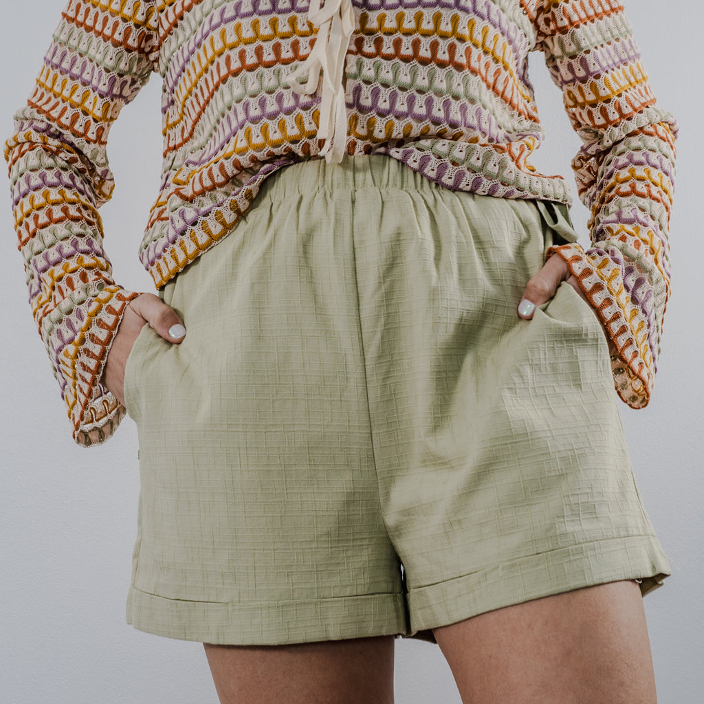 High-waisted sage green crochet shorts with a functional drawstring on either side of hip to adjust waist. Model's hands are partially in pockets to show item has pockets.