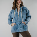 Model wearing a medium-wash denim-style hoodie with a relaxed oversized fit and adjustable hood strings. Hands are in the kangaroo front hoodie pocket.