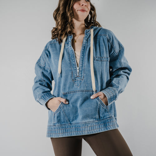 Model wearing a medium-wash denim-style hoodie with a relaxed oversized fit and adjustable hood strings. Hands are in the kangaroo front hoodie pocket.
