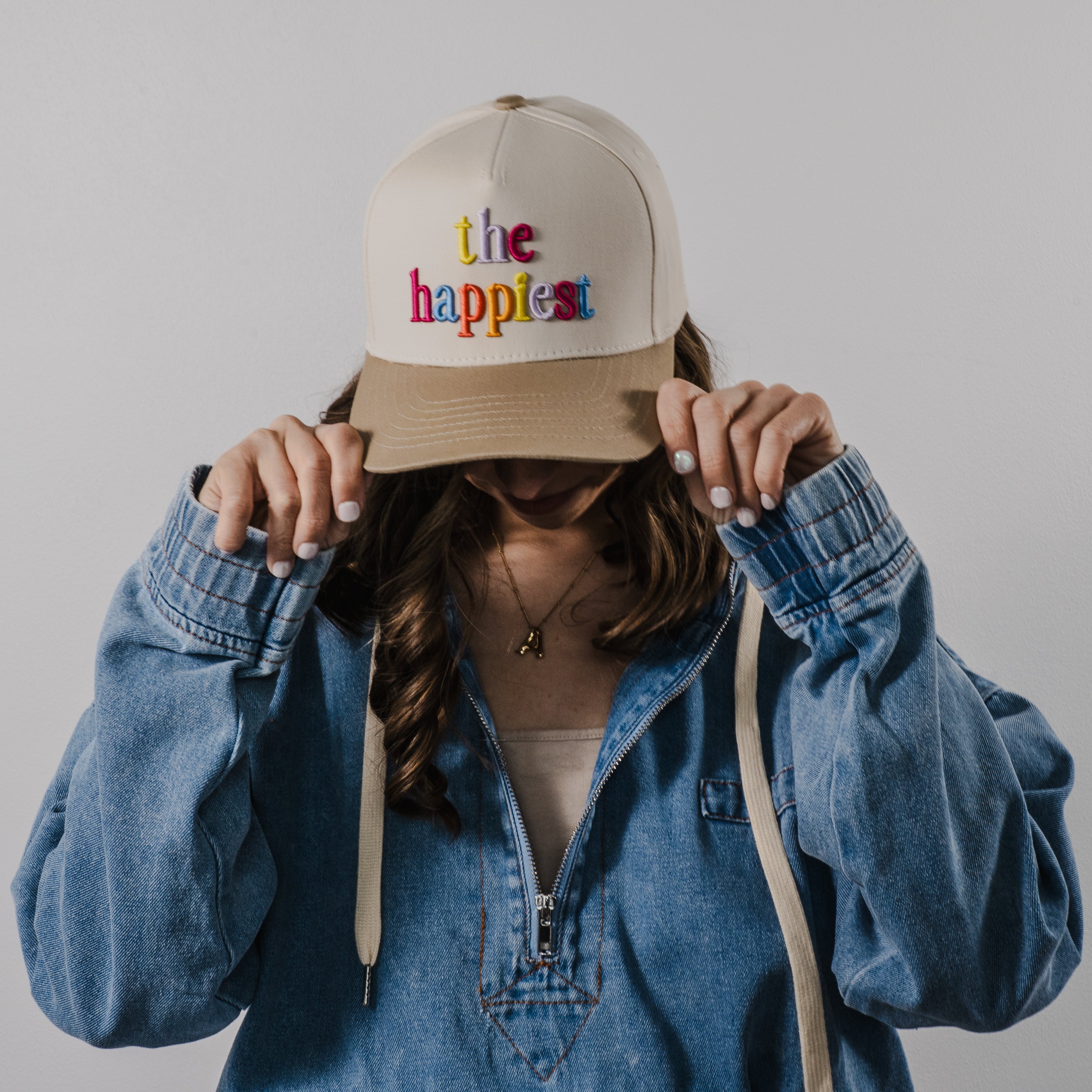 Photography of an unidentified person pulling down a beige baseball cap embroidered with colorful "the happiest" text over their face, wearing a light blue denim jacket with a half-zip and a gold initial A necklace against a plain white indoor wall