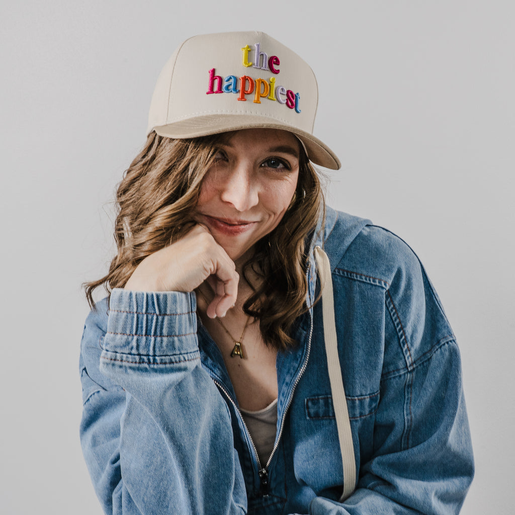A woman in a studio setting wearing a beige baseball cap embroidered with colorful "the happiest" lettering, a blue denim jacket, and a gold initial A pendant necklace