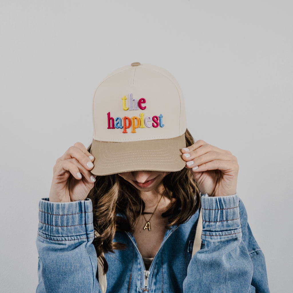 A person with wavy brown hair wearing a blue denim jacket and a gold initial A necklace adjusting a beige baseball cap embroidered with the phrase the happiest in colorful rainbow letters against a plain white background