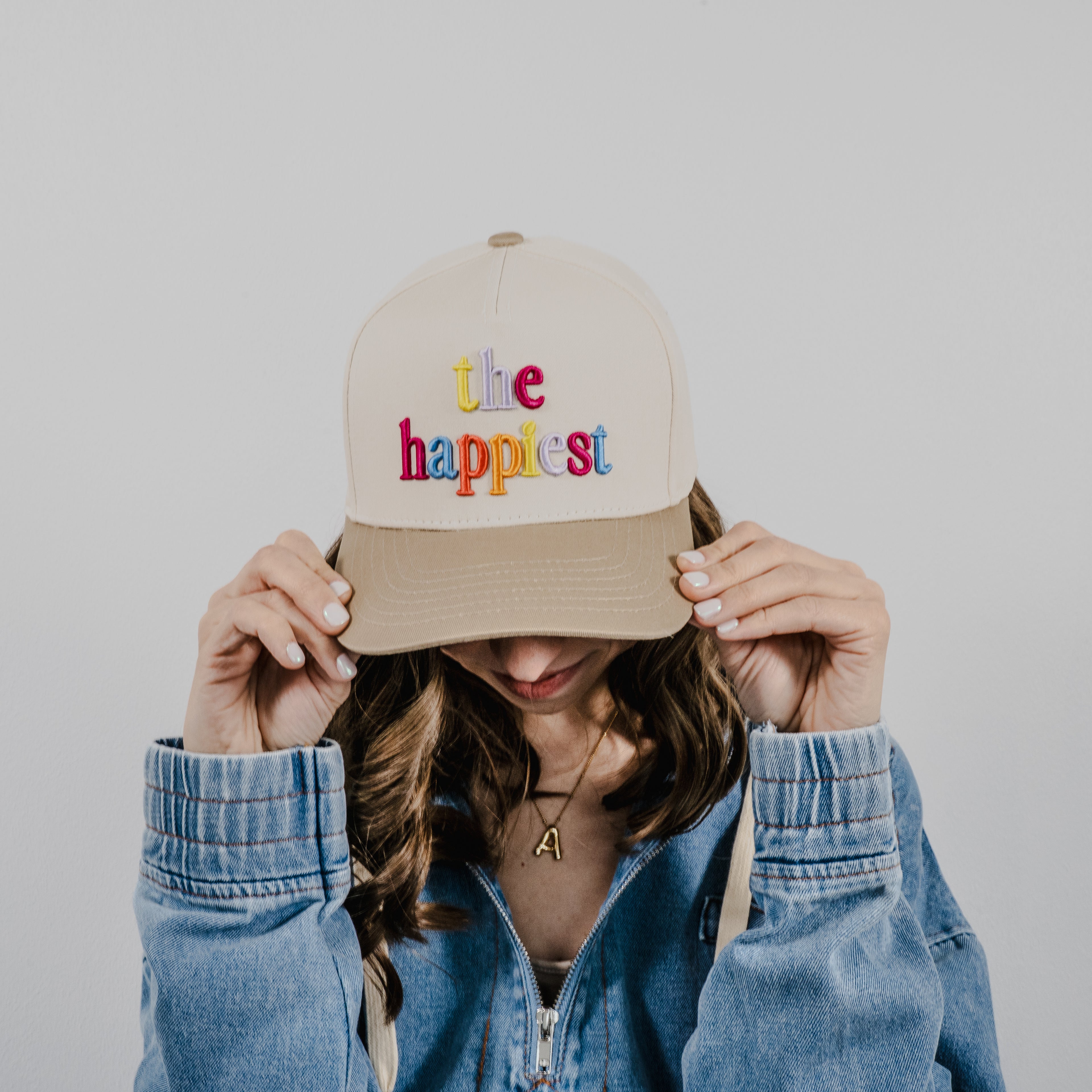 A person with wavy brown hair wearing a blue denim jacket and a gold initial A necklace adjusting a beige baseball cap embroidered with the phrase the happiest in colorful rainbow letters against a plain white background