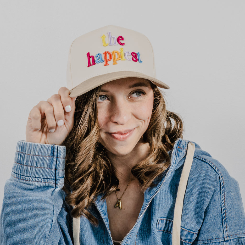 A smiling woman with wavy brown hair holding the brim of her beige hat embroidered with colorful 'the happiest' text, wearing a blue denim jacket and a gold initial A pendant necklace