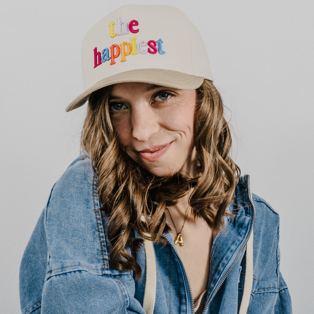 Photograph of a smiling woman with curly brown hair and blue eyes in an indoor studio, wearing a blue denim jacket and a beige baseball cap embroidered with colorful "the happiest" text, showing a golden number 4 pendant on her necklace