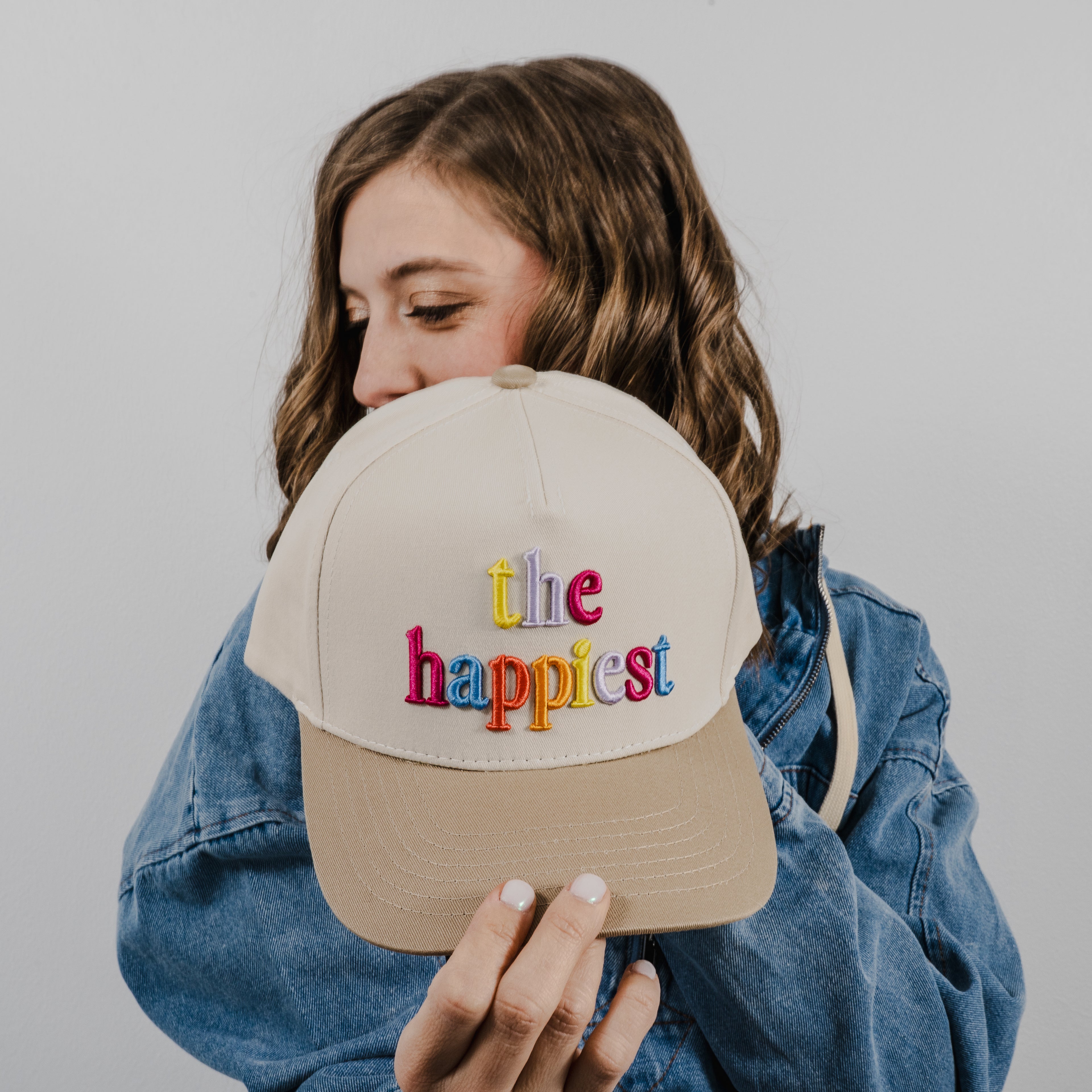 A young woman with wavy brown hair wearing a denim jacket holds a beige and tan baseball cap featuring vibrant multi-colored embroidered text spelling 'the happiest'