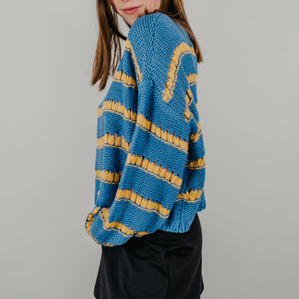 Close-up of the 'Ode to Gloria' sweater sleeve showing the alternating royal blue and golden yellow knit stripes.