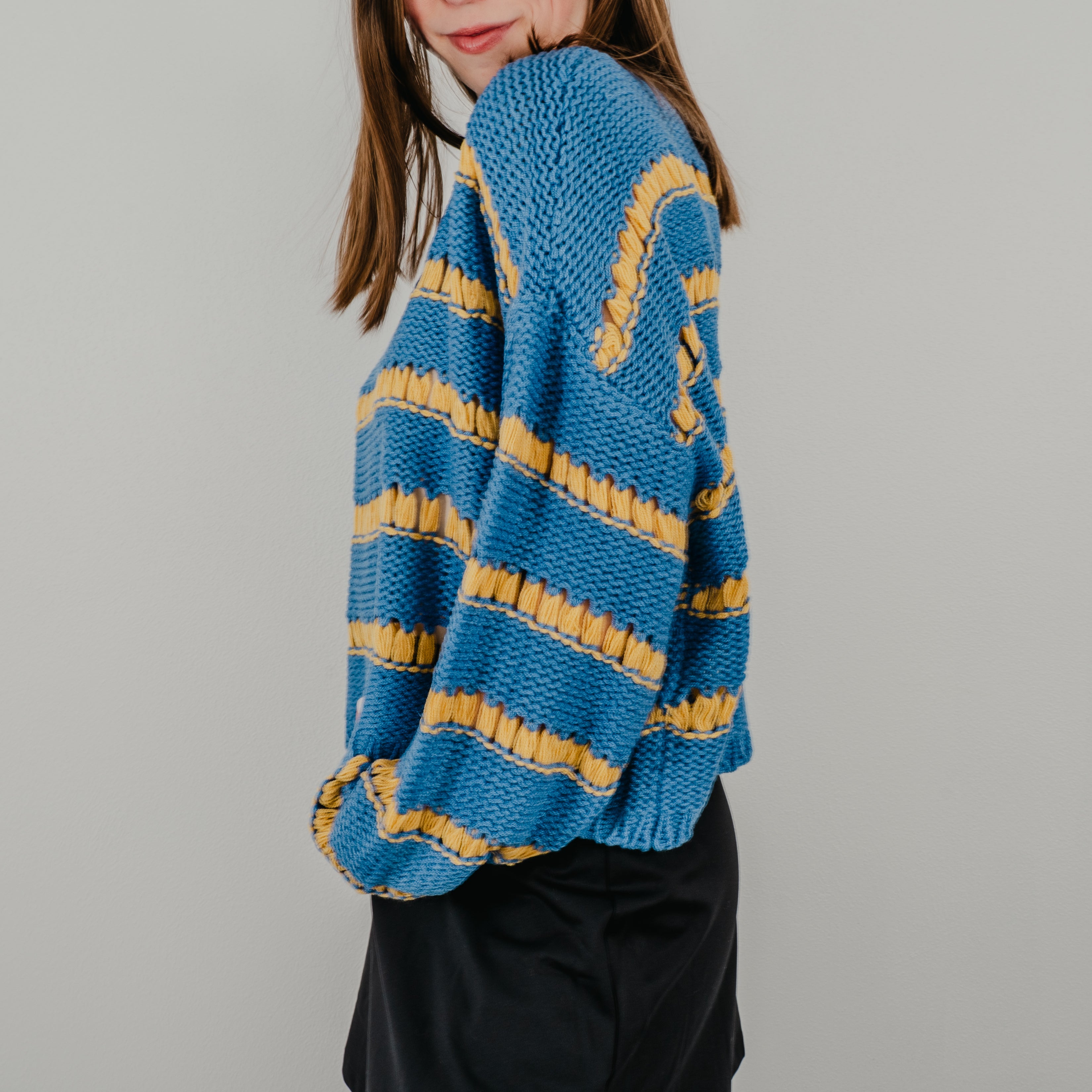 Close-up of the 'Ode to Gloria' sweater sleeve showing the alternating royal blue and golden yellow knit stripes.