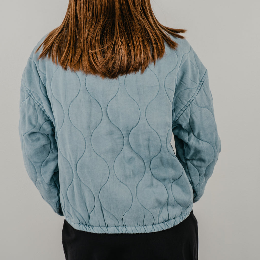 A back view of the Bow Front Quilted Jacket in dust blue showing the curved detailing.
