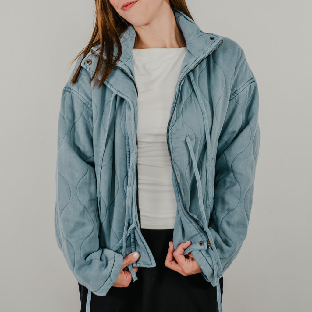 An unzipped view of the lightweight quilted jacket in dusty blue, featuring dangling tie closures down the front and showing optional snap buttons.