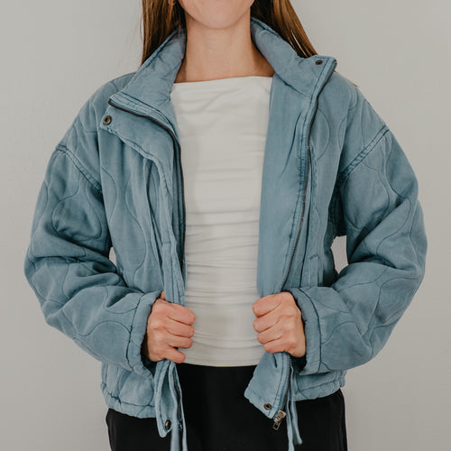 An unzipped view of the lightweight quilted jacket in dusty blue, featuring dangling tie closures down the front and showing optional snap buttons.