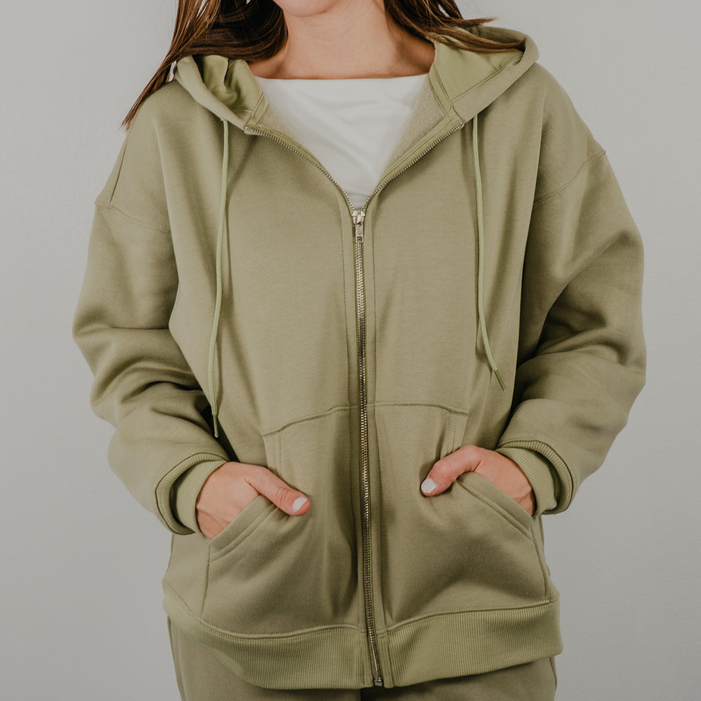 Oversized zip-up hoodie in a premium heavy-weight fleece, featuring a silver front zipper and deep side pockets. Color is a muted, sage green.