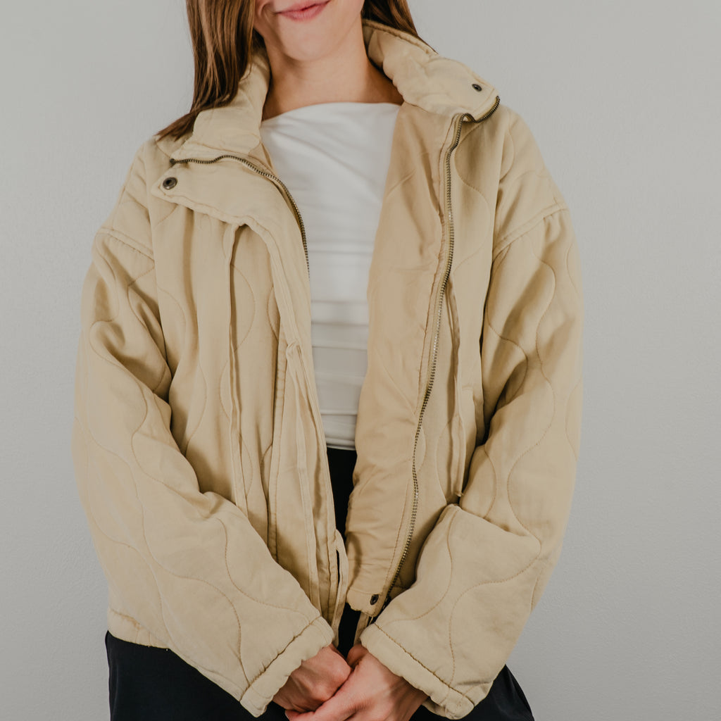An unzipped view of the lightweight quilted jacket in oatmeal, a light tan, featuring dangling tie closures down the front and showing optional snap buttons.