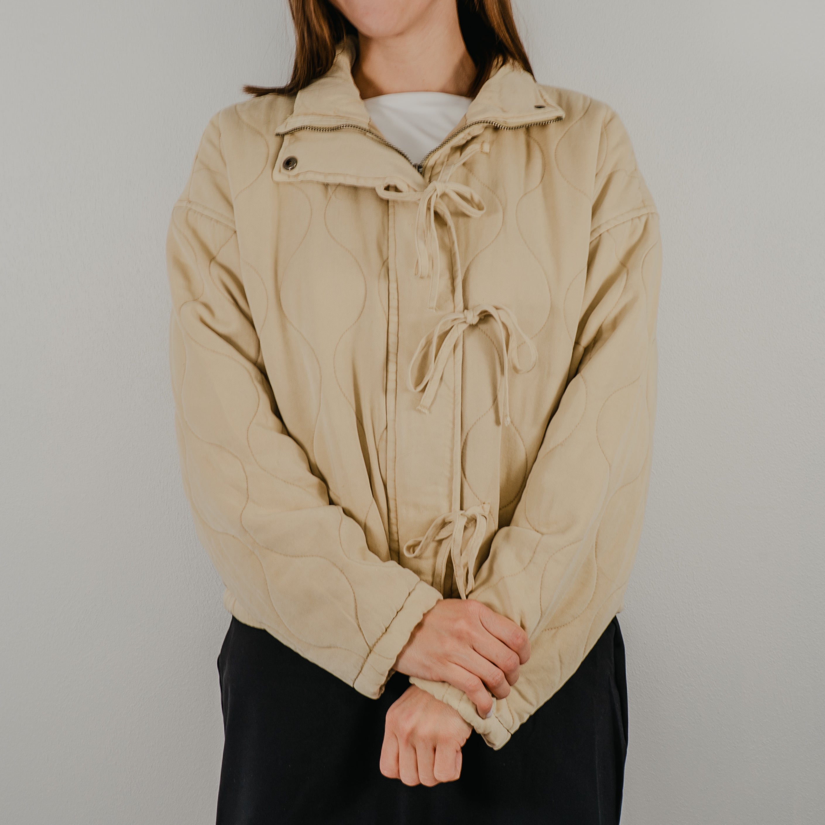 Lightweight quilted jacket in oatmeal, a light tan, featuring delicate bow-tie closures down the front and a collar.