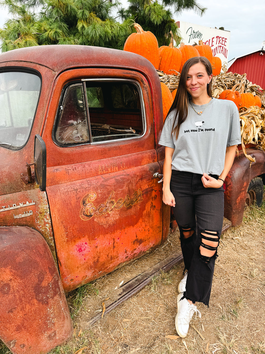 A woman stands outdoors next to a rusted orange vintage pickup truck loaded with pumpkins. She is wearing a light gray graphic T-shirt with the text 'SO YOU'RE YOUNG', black ripped jeans, and white sneakers.
