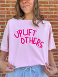 Person wearing a pink t-shirt with 'Uplift Others' text in front of a brick wall.