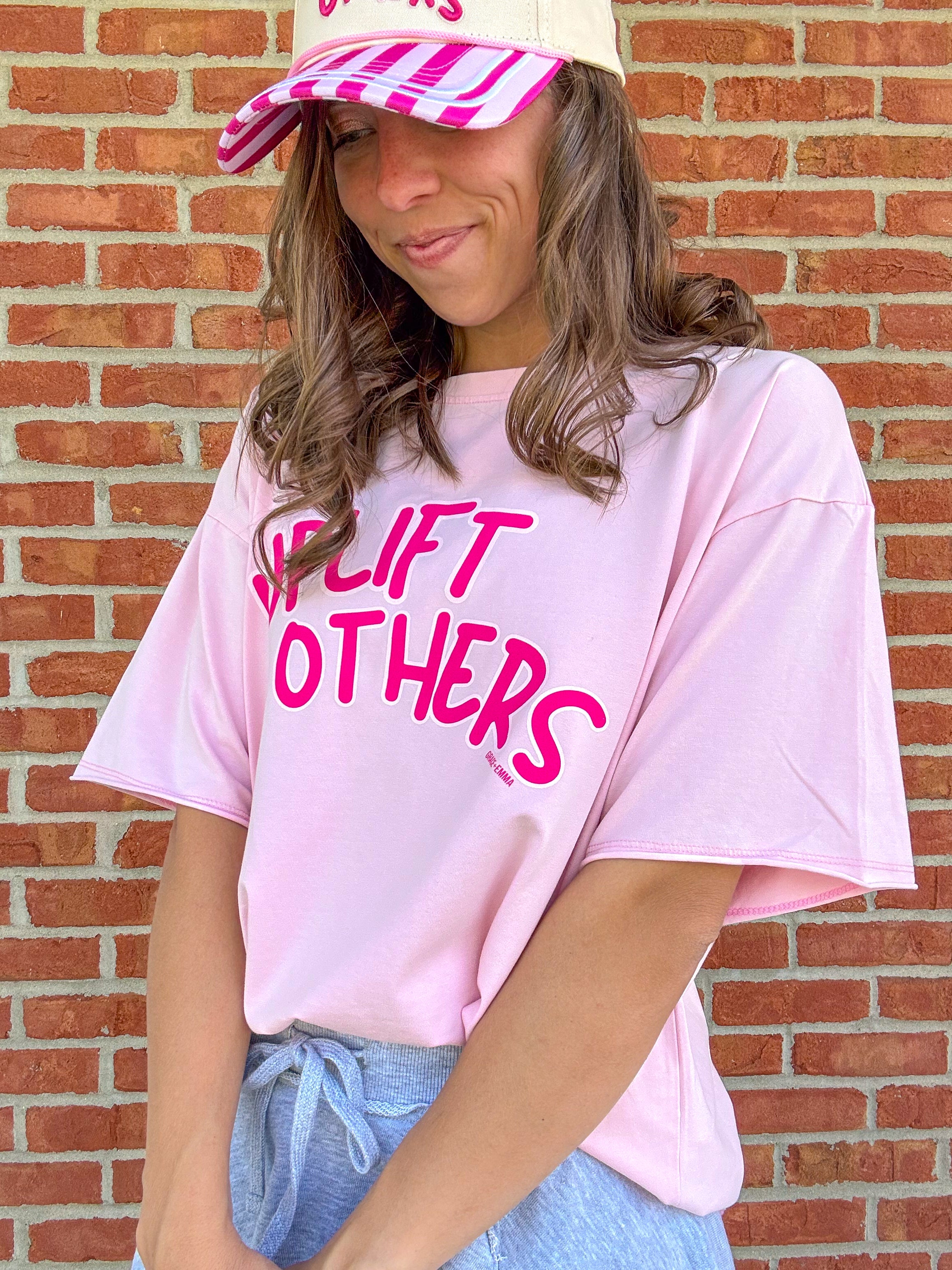Person wearing a pink 'LIFT OTHERS' t-shirt and striped cap against a brick wall.