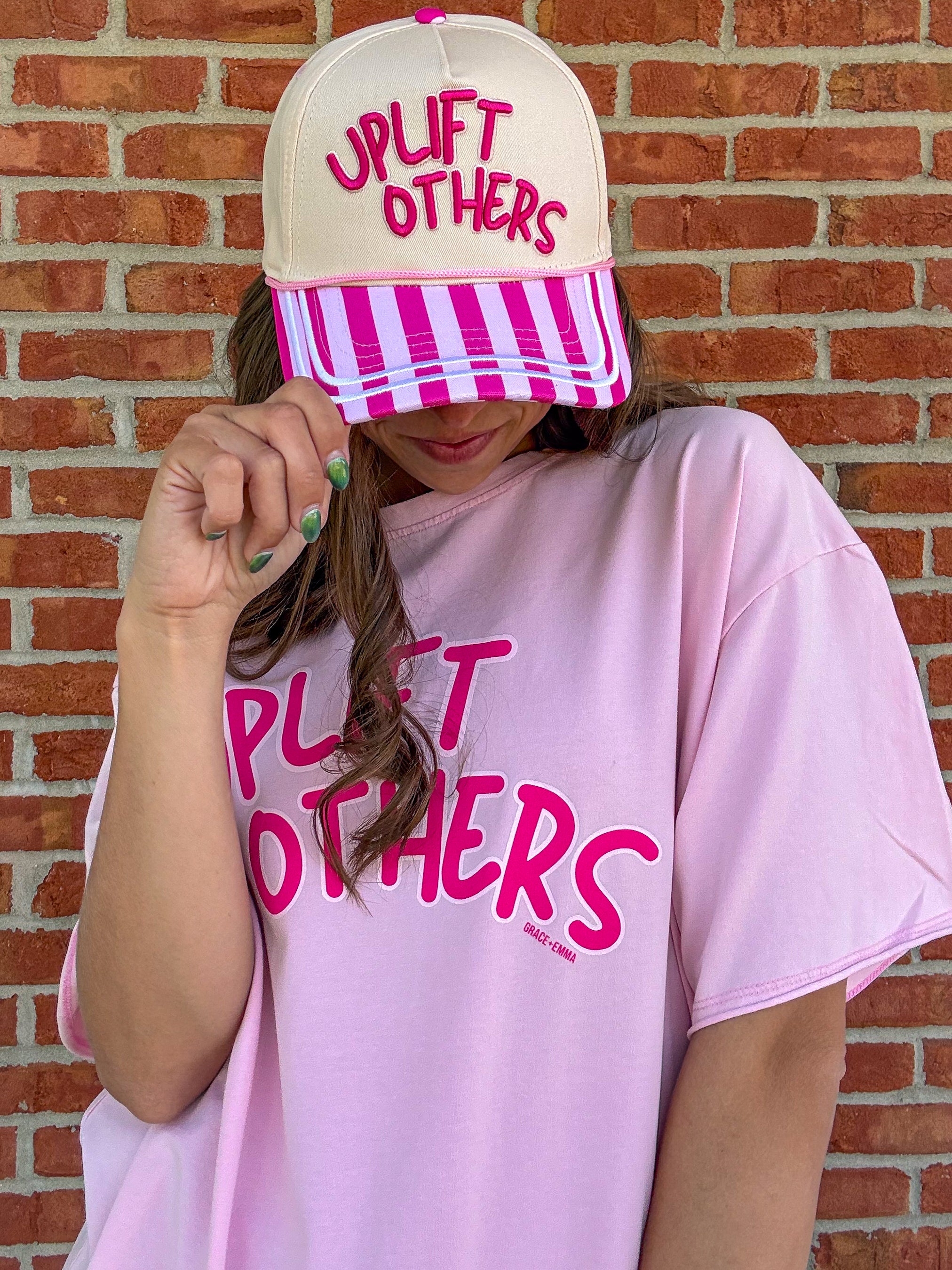 Person wearing a pink 'Uplift Others' t-shirt and cap against a brick wall.