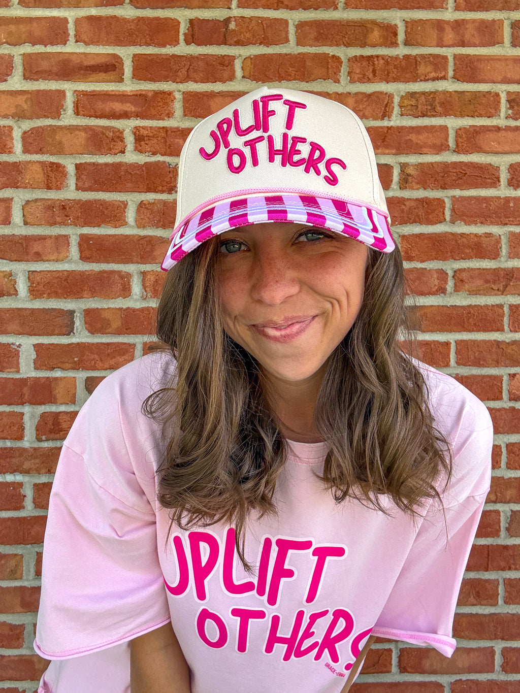 Person wearing a pink and white checkered cap and matching shirt with 'Uplift Others' text in front of a brick wall.