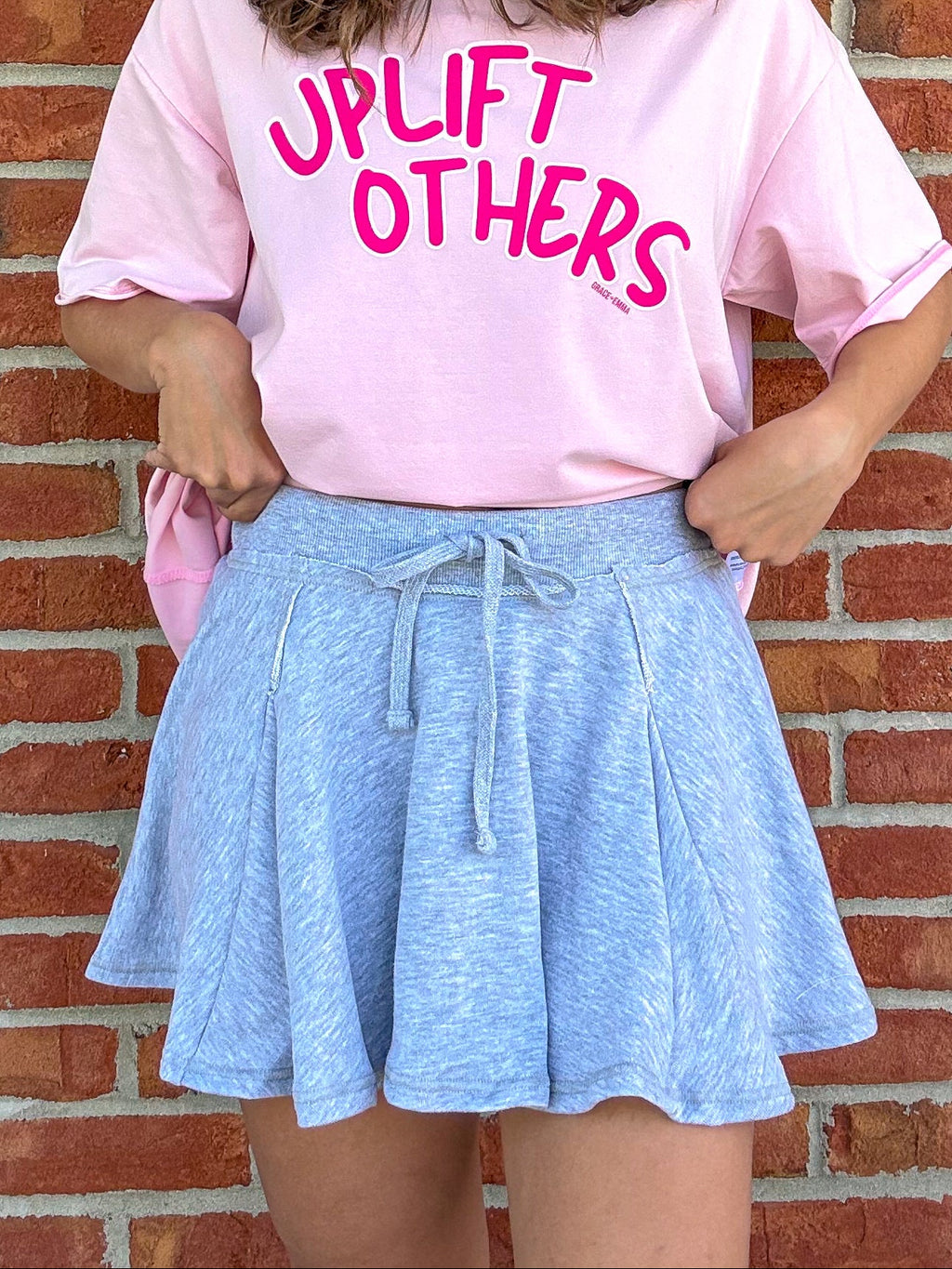 Person wearing a pink 'Uplift Others' shirt and cap against a brick wall.
