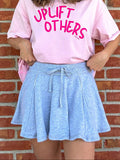 Person wearing a pink 'Uplift Others' shirt and cap against a brick wall.
