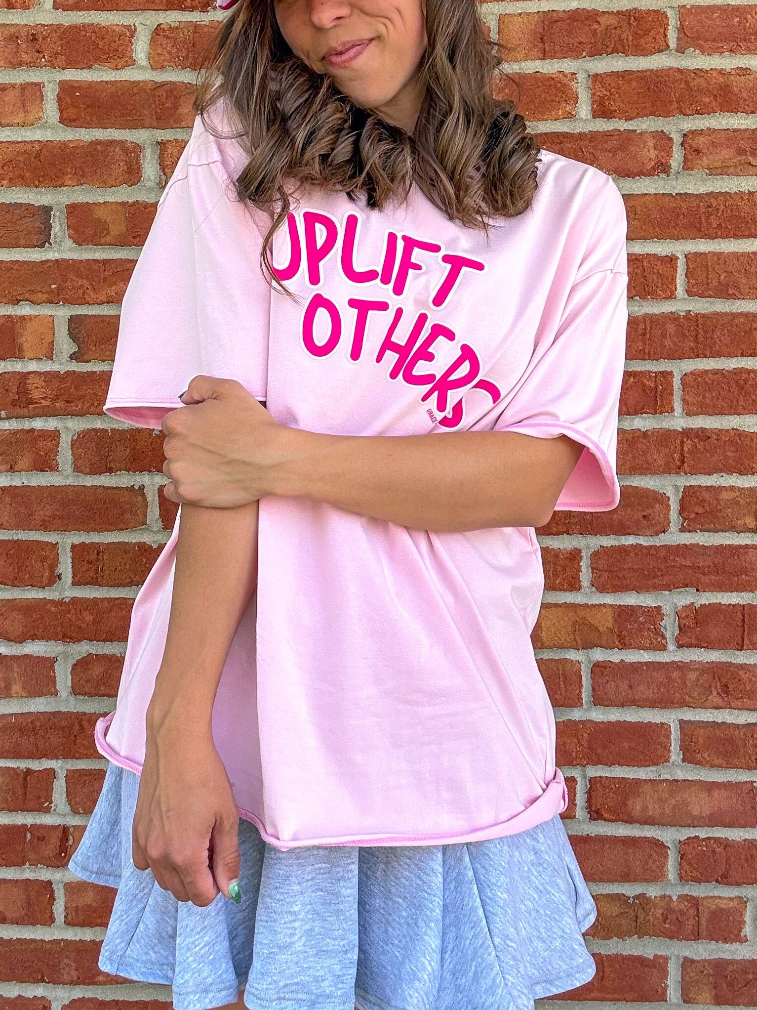 Person wearing a pink 'Uplift Others' t-shirt and cap against a brick wall.