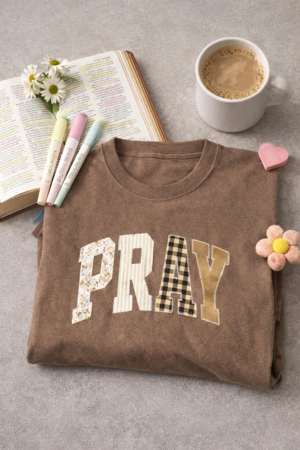 A brown Pray Embroidered Tee neatly folded and resting on a stone surface next to a ceramic coffee mug and an open book, creating a cozy morning aesthetic.