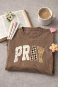 A brown Pray Embroidered Tee neatly folded and resting on a stone surface next to a ceramic coffee mug and an open book, creating a cozy morning aesthetic.